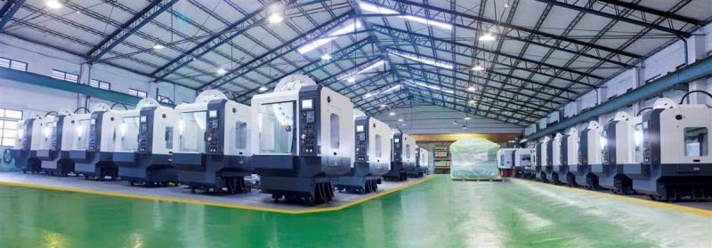 En rymlig industrianläggning inrymmer rader av stora, grå CNC-maskiner, med högt i tak och stark belysning. Golvet är grönmålat med gulkantade gånggångar.