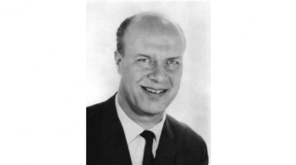 Svartvitt foto av en leende skallig man i mörk kostym och slips.