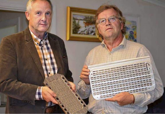 Två män i ledig klädsel håller metallföremål som liknar delar av en mekanisk anordning när de står inomhus.