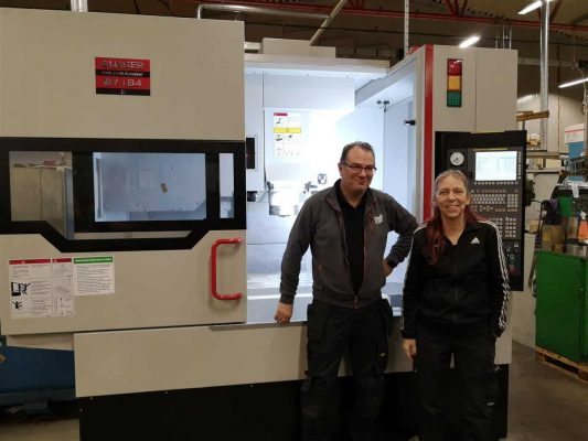 Två personer som står framför en industrimaskin i en verkstad. Maskinen har en digital kontrollpanel och är omgiven av olika verktyg och utrustning.