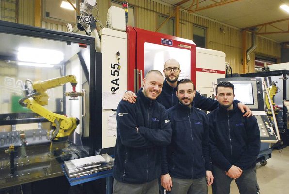 Fyra män i mörka jackor står tillsammans i en tillverkningsanläggning och poserar framför industriutrustning och en robotarm.